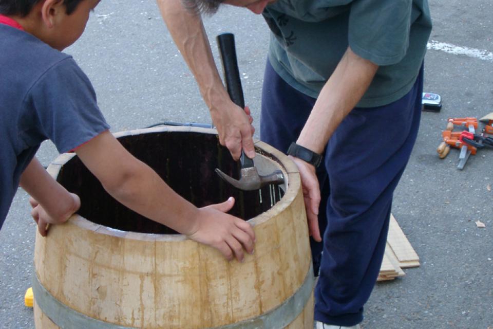 Two taiko members hold down the barrel as they reinforce the rim with a hammer